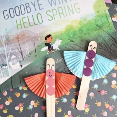 A children's book titled Goodbye Winter, Hello Spring is displayed with two cupcake liner butterfly crafts made from popsicle sticks, cupcake liners, and paper circles, set on vibrant floral paper. Text reads @GLUEDTOMYCRAFTS.