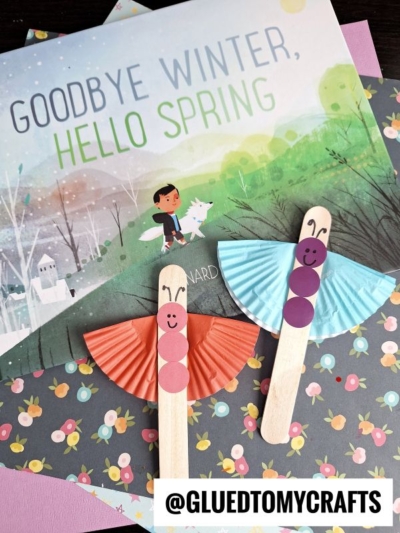 A children's book titled Goodbye Winter, Hello Spring is displayed with two cupcake liner butterfly crafts made from popsicle sticks, cupcake liners, and paper circles, set on vibrant floral paper. Text reads @GLUEDTOMYCRAFTS.