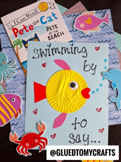 A handmade card with a yellow cupcake liner fish and red fins reads “Swimming by to say…” Surrounded by ocean-themed paper cutouts and the “Pete the Cat: Pete at the Beach” book—perfect for summer craft ideas. @GLUEDTOMYCRAFTS watermark at the bottom.