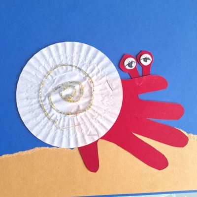 A snail craft made from a red paper handprint for the body and a white cupcake liner for the shell, with googly eyes on red stalks, on a blue and tan background. The username @GLUEDTOMYCRAFTS is at the bottom.