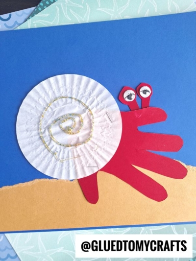 A snail craft made from a red paper handprint for the body and a white cupcake liner for the shell, with googly eyes on red stalks, on a blue and tan background. The username @GLUEDTOMYCRAFTS is at the bottom.