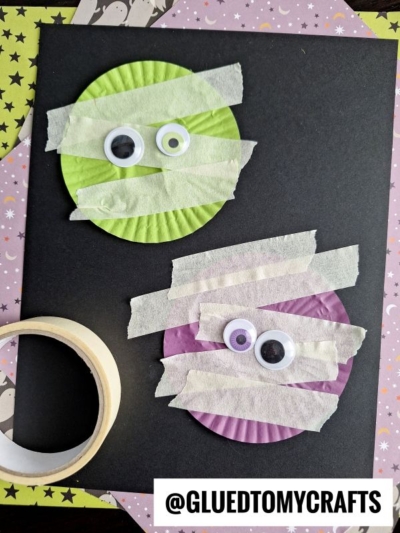 Two cupcake liners, one green and one purple, are decorated as mummies with strips of white tape and googly eyes. They are placed on black paper with a roll of tape nearby. @GLUEDTOMYCRAFTS is written at the bottom.