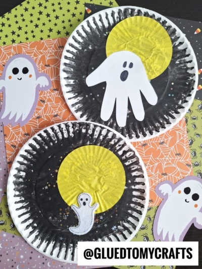 Two Halloween-themed paper plate crafts with black borders, yellow moons, and white ghosts made from cutouts. The plates are on orange and green Halloween-patterned paper with cartoon ghost decorations.