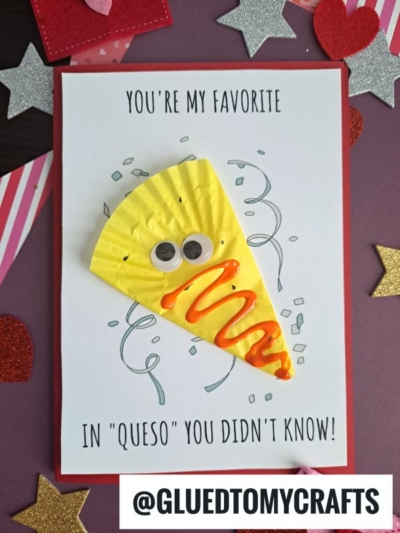 A handmade cupcake liner card crafts a pun saying, “You’re my favorite in ‘queso’ you didn’t know!” with a smiling cheese wedge made from a yellow cupcake liner, googly eyes, and puffy orange writing, surrounded by star and heart cutouts.