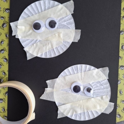 Two paper cupcake liners taped to black paper with masking tape strips, each decorated with two googly eyes to resemble mummies. A roll of tape sits nearby on a Halloween-themed background. @GLUEDTOMYCRAFTS is written at the bottom.