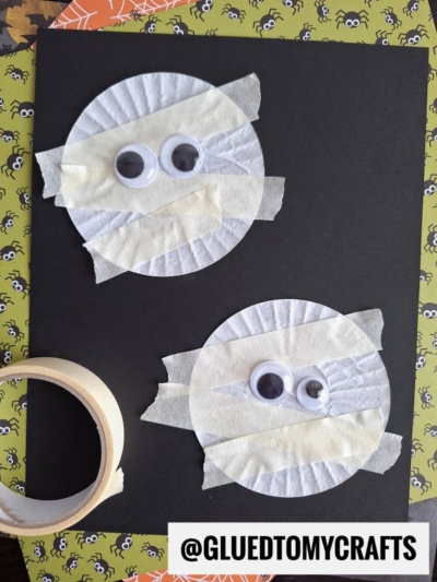 Two paper cupcake liners taped to black paper with masking tape strips, each decorated with two googly eyes to resemble mummies. A roll of tape sits nearby on a Halloween-themed background. @GLUEDTOMYCRAFTS is written at the bottom.