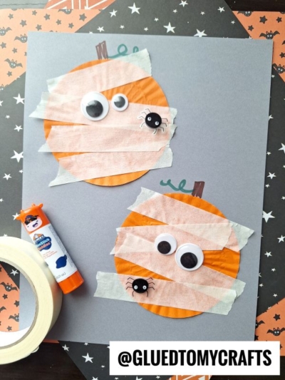 Two orange paper circles decorated as mummies with strips of white tape, googly eyes, and small black spider stickers are glued to gray paper. A glue stick and roll of tape are beside them. Halloween-themed paper is in the background.