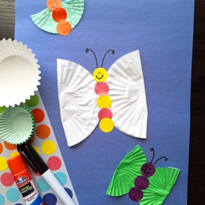 A craft project featuring paper butterflies made from cupcake liners and colorful stickers on a blue background, with a glue stick, marker, and sticker sheet nearby. Text reads @GLUEDTOMYCRAFTS.