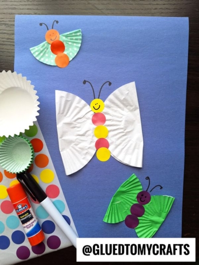 A craft project featuring paper butterflies made from cupcake liners and colorful stickers on a blue background, with a glue stick, marker, and sticker sheet nearby. Text reads @GLUEDTOMYCRAFTS.