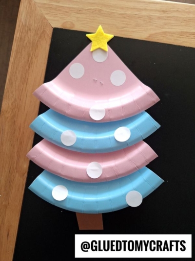 A Christmas tree craft made from pink and blue paper plate pieces with white dot decorations, a yellow star on top, and a brown rectangle for the trunk. Handle is visible in the background. Text: @GLUEDTOMYCRAFTS.