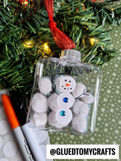 A clear ornament filled with white pom-poms and glitter, shaped like a snowman, hangs from a Christmas tree. A red ribbon is tied to the top, making it perfect for coding label crafts this holiday season.