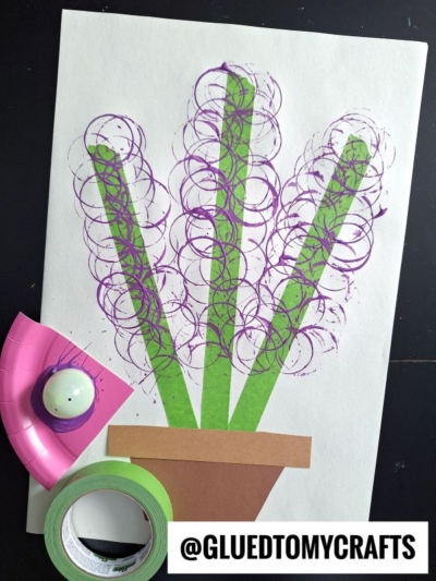 A paper craft of a potted plant with green stems and purple circular flowers, made using colored tape, paint, and the Plastic Egg Stamp Crafts technique. A paint-covered cup, pink tape, green tape, and @GLUEDTOMYCRAFTS are also visible.