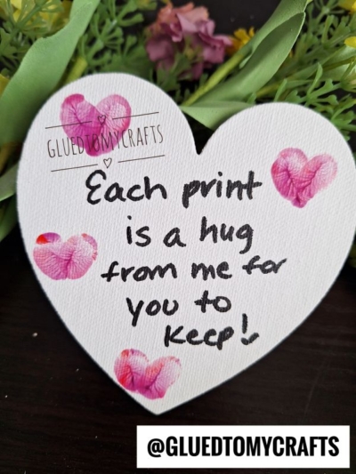 A heart-shaped paper with pink fingerprints and the handwritten message, “Each print is a hug from me for you to keep!” rests on a dark surface with green leaves and flowers—a sweet thumbprint craft idea for spring. @GLUEDTOMYCRAFTS is at the bottom.