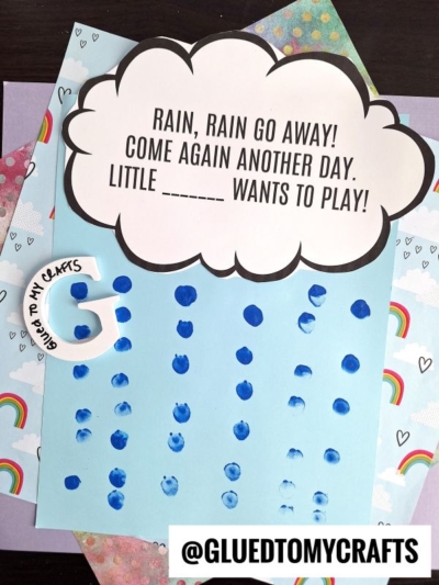 A paper craft features a large cloud cutout with the text RAIN, RAIN GO AWAY! COME AGAIN ANOTHER DAY. LITTLE _____ WANTS TO PLAY! Blue fingerprints form raindrops on blue paper below, surrounded by rainbow-themed paper.
