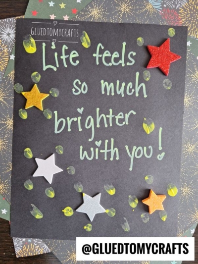 A black paper with the handwritten message Life feels so much brighter with you! decorated with colorful star cutouts and yellow thumbprints, makes a cheerful THUMBPRINT CRAFTS FOR KIDS idea. @GLUEDTOMYCRAFTS is at the bottom.