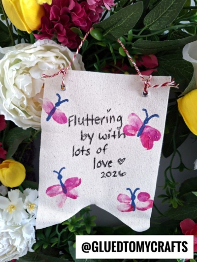 A small fabric banner decorated with pink and blue butterflies and the handwritten message Fluttering by with lots of love ♥ 2024. Perfect for THUMBPRINT CRAFT IDEAS FOR SPRING, it hangs among colorful flowers. @GLUEDTOMYCRAFTS is labeled at the bottom.