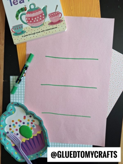A pink sheet of paper with three green lines, a green marker, a cupcake-shaped plate with a spoon and ball, patterned papers, and a childrens tea party book, all arranged on a table. @GLUEDTOMYCRAFTS is at the bottom.