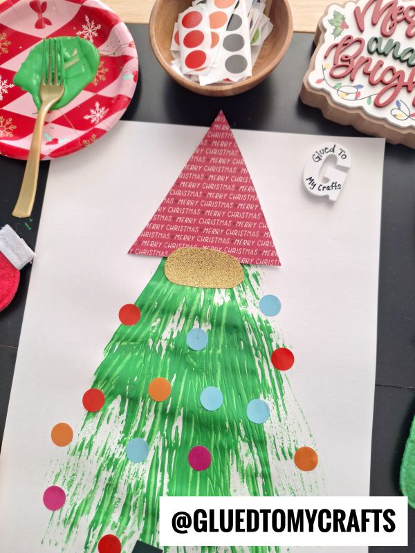 A handmade Christmas tree craft on white paper using green paint, a red triangle for the top, and colorful dot stickers as ornaments. Supplies and tools surround the artwork. @GLUEDTOMYCRAFTS is written at the bottom.