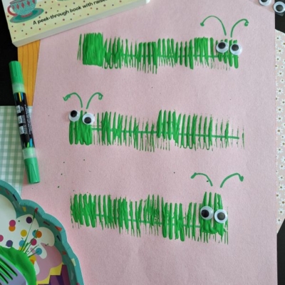 A pink paper displays three green caterpillars made from marker lines and googly eyes. Surrounding the paper are a book, markers, googly eyes, cupcake liners, and plastic utensils. Text reads @GLUEDTOMYCRAFTS.