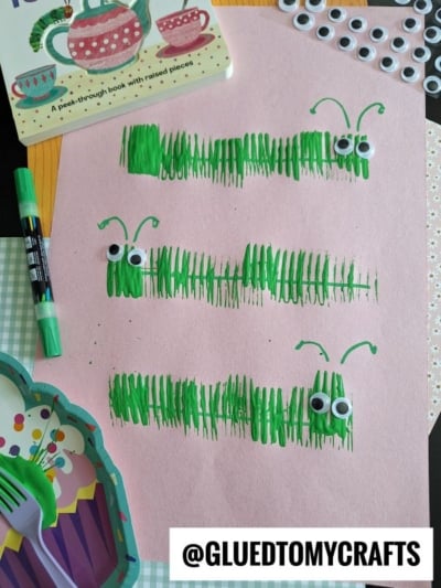 A pink paper displays three green caterpillars made from marker lines and googly eyes. Surrounding the paper are a book, markers, googly eyes, cupcake liners, and plastic utensils. Text reads @GLUEDTOMYCRAFTS.