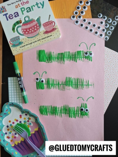 A pink sheet of paper features three green caterpillars made from marker lines and googly eyes. Surrounding items include a childrens tea party book, a marker, craft paper, googly eyes, and a cupcake-shaped plate.
