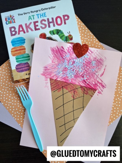 A children’s book titled “The Very Hungry Caterpillar at the Bakeshop” sits next to a handmade ice cream craft with pink paint, a red glitter heart, and blue sprinkles on paper. A blue fork and patterned paper are nearby.