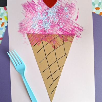A handmade ice cream cone craft on paper, featuring a brown cone with black lines, pink textured ice cream topped with glitter and a red heart, lying next to a blue plastic fork. @GLUEDTOMYCRAFTS is written at the bottom.