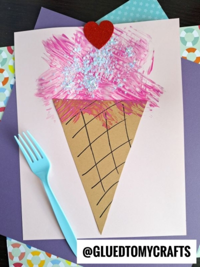 A handmade ice cream cone craft on paper, featuring a brown cone with black lines, pink textured ice cream topped with glitter and a red heart, lying next to a blue plastic fork. @GLUEDTOMYCRAFTS is written at the bottom.