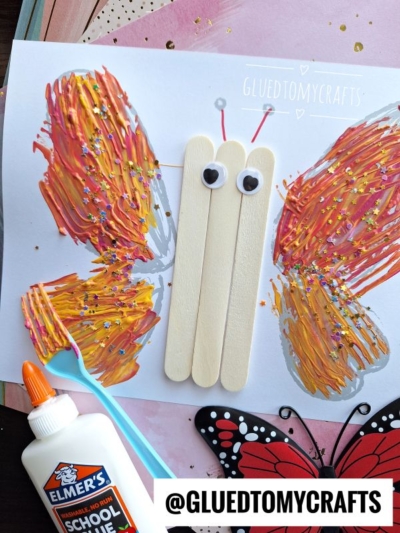 A butterfly craft made with painted wings using red, orange, and yellow streaks, two wooden sticks for the body, googly eyes, and glitter, shown with glue and a plastic fork on the side.