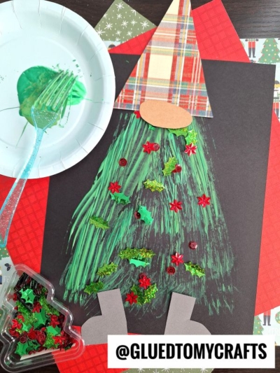 A Christmas gnome craft made with a plaid paper hat, oval nose, green painted beard created with a fork, gray boots, and decorated with sequins and glitter shapes. A plate with green paint and a plastic fork is nearby.