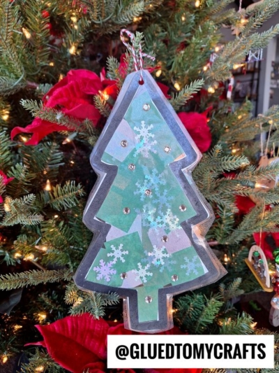A handmade stained glass Christmas tree ornament with snowflake designs and gems hangs on a decorated tree adorned with red poinsettias and lights. @GLUEDTOMYCRAFTS is written at the bottom of the image.