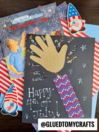 A handmade Fourth of July card with a gold handprint as a firework, surrounded by glitter stars, sits on a table with patriotic stickers and a book titled The Night Before the Fourth of July—perfect for 4th of July keepsakes.