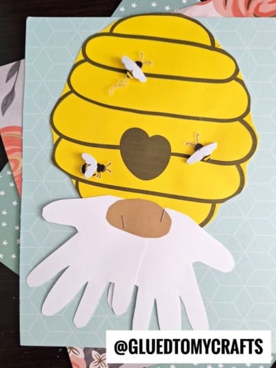 A charming Paper Beehive Gnome craft featuring a yellow beehive with bees, a heart-shaped entrance, and a white handprint beard, displayed on patterned paper with the text @GLUEDTOMYCRAFTS. Perfect for any bee gnome lover!.