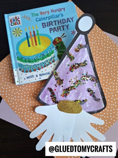 A children’s book, “The Very Hungry Caterpillar’s Birthday Party,” sits on orange polka dot paper next to a handprint birthday gnome sporting a purple party hat decorated with glitter and confetti. @GLUEDTOMYCRAFTS is at the bottom.