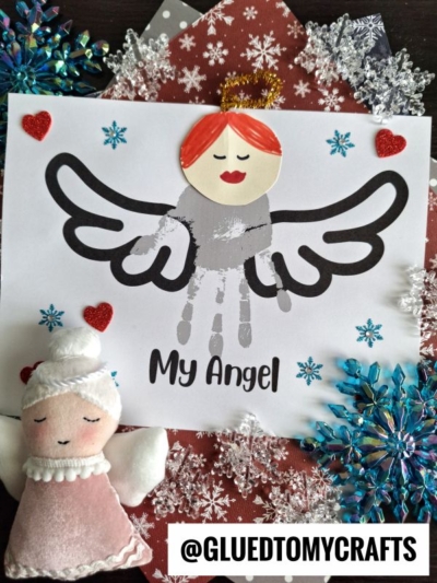A charming handprint angel craft features a painted handprint for the body and wings, with a round face, red hair, and halo. Snowflakes and a plush angel doll accent the background. Text reads My Angel and @GLUEDTOMYCRAFTS.