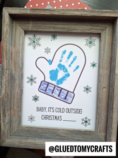 A framed artwork features a large mitten outline with a blue handprint inside, surrounded by snowflake designs. Text below reads, BABY, ITS COLD OUTSIDE! CHRISTMAS Mitten Keepsake Printable. The frame is rustic wood.