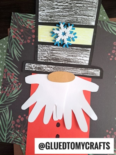 A handmade snowman craft with a hat, paper snowflake, orange nose, and a white handprint beard on festive paper—perfect for pairing with your favorite nutcracker gnome decor. @GLUEDTOMYCRAFTS is written at the bottom.