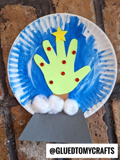 A paper plate craft featuring a blue painted background, a cut-out green handprint snowman snow globe with red gems and a yellow star, white cotton balls at the base, a gray stand, and @GLUEDTOMYCRAFTS written at the bottom.
