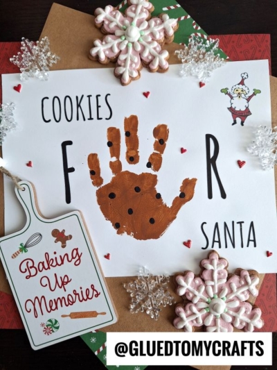 A festive craft featuring a child’s brown handprint with black dots as cookies for Santa, surrounded by snowflake cookies, a “Baking Up Memories” sign, and Christmas-themed paper.