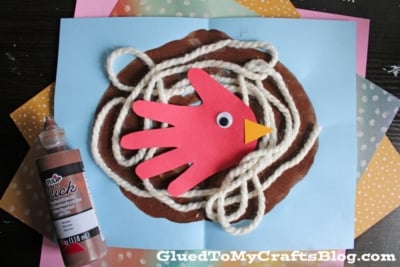 A childrens craft shows a red paper handprint bird with a googly eye and orange beak in a yarn nest on blue paper. Brown paint and a bottle of brown fabric paint are nearby. GluedToMyCraftsBlog.com is at the bottom.