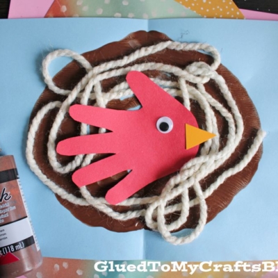 A childrens craft shows a red paper handprint bird with a googly eye and orange beak in a yarn nest on blue paper. Brown paint and a bottle of brown fabric paint are nearby. GluedToMyCraftsBlog.com is at the bottom.