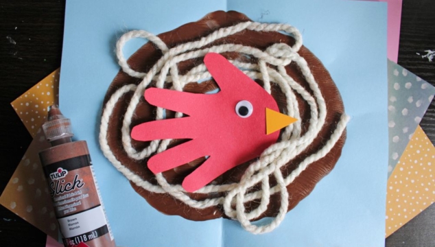 A childrens craft shows a red paper handprint bird with a googly eye and orange beak in a yarn nest on blue paper. Brown paint and a bottle of brown fabric paint are nearby. GluedToMyCraftsBlog.com is at the bottom.