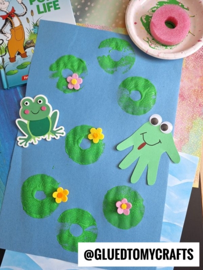 Blue paper with green lily pad stamps, pink and yellow flower embellishments, a paper frog, and a green handprint frog with googly eyes. Art supplies and a book are visible in the background.