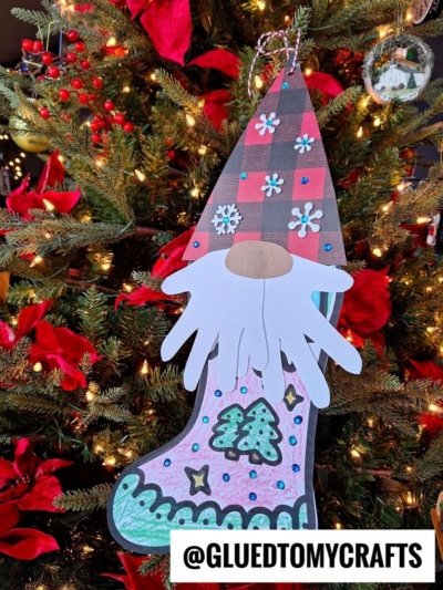 A colorful gnome stocking ornament, shaped like a paper gnome with trees, snowflakes, and patterns, hangs on a Christmas tree adorned with red flowers and lights. A tag reads @GLUEDTOMYCRAFTS.