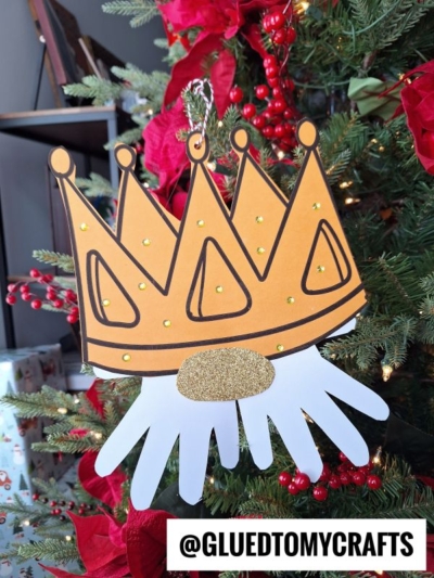 A handmade Christmas ornament shaped like a gold crown with jewel accents, attached to a white handprint cutout, resembles a charming handprint king gnome. The ornament hangs on a decorated tree and is labeled @GLUEDTOMYCRAFTS at the bottom.