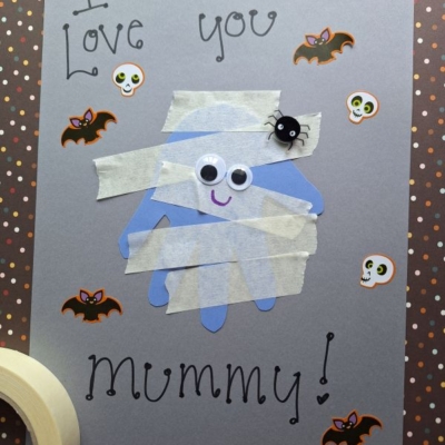 A handmade mummy card with “I love you Mummy!” written on gray paper. The mummy is made from a blue handprint, tape strips, googly eyes, and a small spider. Decorated with stickers of bats and skulls.