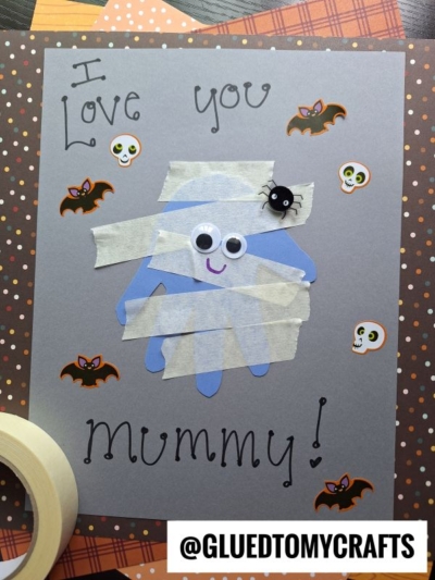 A handmade mummy card with “I love you Mummy!” written on gray paper. The mummy is made from a blue handprint, tape strips, googly eyes, and a small spider. Decorated with stickers of bats and skulls.