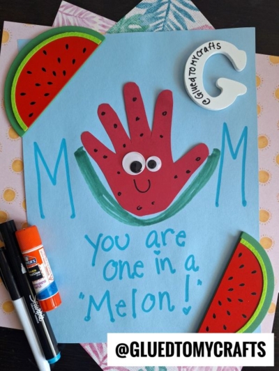 A handmade card with MOM in blue, featuring a red handprint decorated as a smiling watermelon slice with googly eyes. Perfect for mother's day keepsakes, it says You are one in a Melon! and is surrounded by watermelon slices and markers.