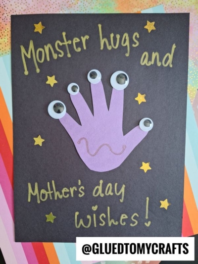 A handmade Mother's Day card features a purple handprint with googly eyes, a squiggly line, gold star stickers, and the text “Monster hugs and Mother’s day wishes!” on black paper—perfect for cherished mother's day keepsakes.