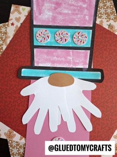A holiday craft featuring a nutcracker gnome made of paper, with a white handprint beard, pink hat decorated with candy images, and a brown oval nose. Background includes patterned Christmas paper. @GLUETOMYCRAFTS.